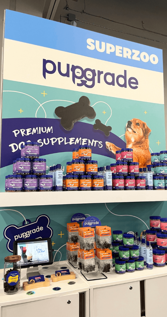 A Pupgrade booth at SuperZoo 2024 displaying various dog supplements and treats. The booth features a large banner with the Pupgrade logo and the words "PREMIUM DOG SUPPLEMENTS." There are shelves stocked with colorful jars of supplements and treats, as well as a touchscreen device and other promotional materials.