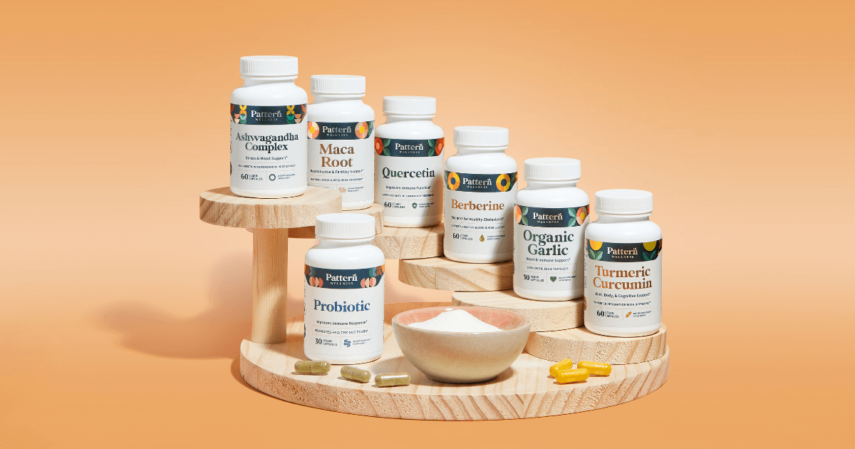 Close-up shot of Pattern brand vitamins including Ashwagandha, Maca Root, Quercetin, Berberine, Garlic, and Turmeric, displayed on a light wood tiered stand against a peach background.